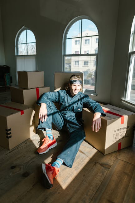 A young man with light skin and long hair tied back, wearing a dark blue jumpsuit, sits on a wooden floor inside a room with large arched windows and white walls. He is smiling and relaxed, leaning against several cardboard boxes sealed with red tape, which are stored on the floor and near the window. The boxes vary in size and are likely filled with household items for a home relocation. Natural sunlight streams through the windows, illuminating the scene and casting shadows on the floor. The room appears to be in a residential property, and the image captures a moment in the packing and moving process. This setting exemplifies furniture transport and packing activities performed by a professional removal service such as Man With a Van Stockwell, supporting efficient household move arrangements.
