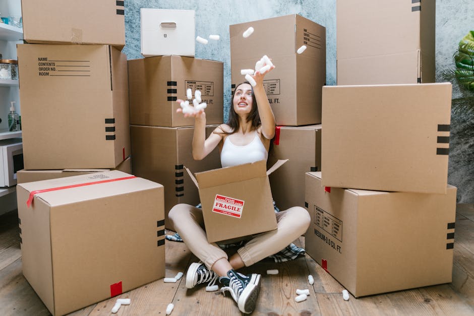 A woman sitting cross-legged on a wooden floor, surrounded by numerous cardboard moving boxes of various sizes, some sealed with red tape and others open with packing materials. She is inside a room with light blue painted walls and a potted plant visible on the right side, suggesting a home environment. The woman is wearing a white tank top, beige pants, and black-and-white sneakers, and she is smiling while tossing pieces of foam packing insert into the air. Several foam packing inserts are scattered around her, and she appears to be engaged in packing or unpacking during a house relocation process. Man With a Van Stockwell, a professional removals service, is involved in packing and moving furniture and boxes, as depicted by the scene of careful handling and storage of household items at the start or end of a home moving project.