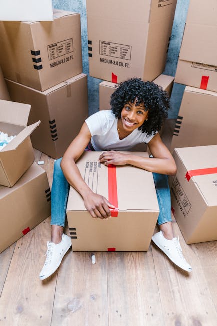 A young man with light skin and long hair tied back, wearing a dark blue jumpsuit, sits on a wooden floor inside a room with large arched windows and white walls. He is smiling and relaxed, leaning against several cardboard boxes sealed with red tape, which are stored on the floor and near the window. The boxes vary in size and are likely filled with household items for a home relocation. Natural sunlight streams through the windows, illuminating the scene and casting shadows on the floor. The room appears to be in a residential property, and the image captures a moment in the packing and moving process. This setting exemplifies furniture transport and packing activities performed by a professional removal service such as Man With a Van Stockwell, supporting efficient household move arrangements.