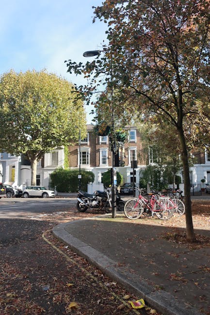 A view of a residential street with a small parking area and sidewalk, captured during daylight hours with clear blue skies. Several bicycles, including a pink children's bike, are parked against a black lamppost near the curb, and a black motorcycle is also parked on the street. In the background, there are white and brick terraced houses with large windows and front gardens, partially obscured by trees with green and autumn-colored leaves. The pavement is lined with fallen leaves, and the scene suggests a calm neighbourhood environment. This image is relevant to house removals and moving services, illustrating the typical setting where a professional removal company such as Man With a Van Stockwell might facilitate furniture transport during a home relocation process.