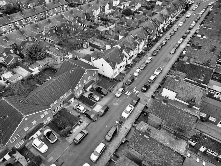 A view of a residential street with a small parking area and sidewalk, captured during daylight hours with clear blue skies. Several bicycles, including a pink children's bike, are parked against a black lamppost near the curb, and a black motorcycle is also parked on the street. In the background, there are white and brick terraced houses with large windows and front gardens, partially obscured by trees with green and autumn-colored leaves. The pavement is lined with fallen leaves, and the scene suggests a calm neighbourhood environment. This image is relevant to house removals and moving services, illustrating the typical setting where a professional removal company such as Man With a Van Stockwell might facilitate furniture transport during a home relocation process.